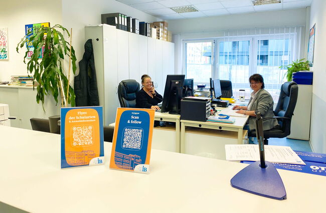 Im Sekretariat arbeiten zwei Frauen an Schreibtischen, während Informationsflyer und QR-Codes im Vordergrund stehen.