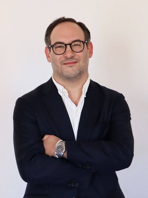 Ein Mann in einem dunklen Anzug und Brille lächelt selbstbewusst mit verschränkten Armen vor einem hellen Hintergrund.