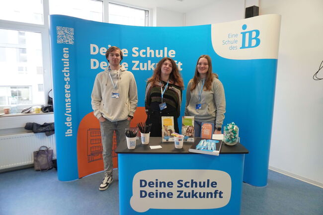 Drei Personen stehen an einem Informationsstand mit dem Slogan "Deine Schule, Deine Zukunft" und Broschüren.