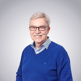 Ein lächelnder Mann in blauem Pullover steht vor einem neutralen Hintergrund und schaut freundlich in die Kamera.