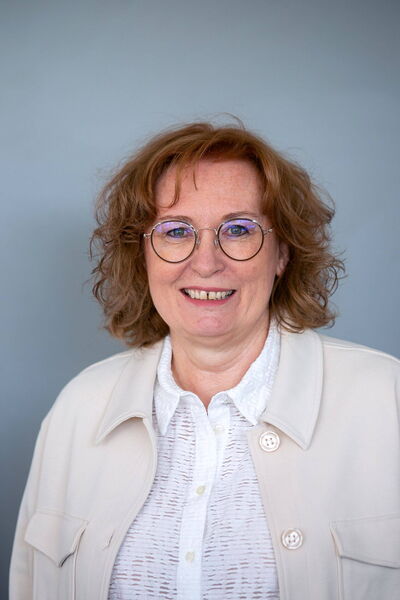 Eine lächelnde Frau mit Brille und lockigem Haar trägt eine weiße Bluse vor einem neutralen Hintergrund.
