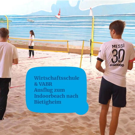 Zwei Personen spielen Beachvolleyball in einer Indoorhalle mit Sandboden und Wandgemälde, während eine dritte Person zuschaut.