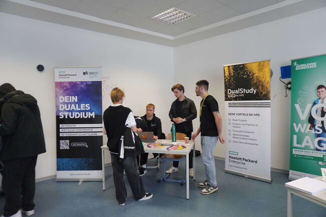 Mehrere junge Männer informieren sich an einem Stand über duale Studienmöglichkeiten bei einer Messe.