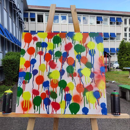 Ein buntes Graffiti-Kunstwerk mit Farbflecken steht auf einer Staffelei vor einem Schulgebäude im Freien.