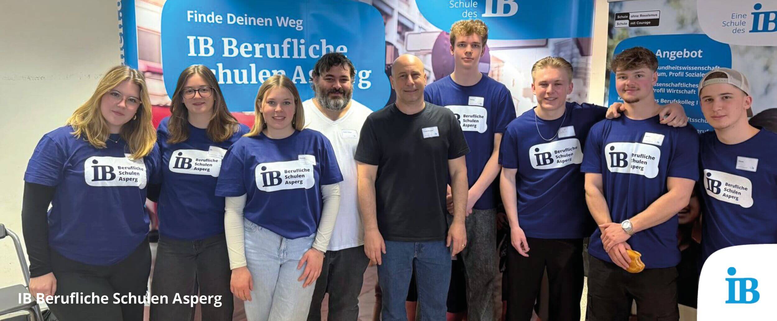 Eine Gruppe von Menschen steht lächelnd vor einem Banner der IB Beruflichen Schulen Asperg während einer Veranstaltung.