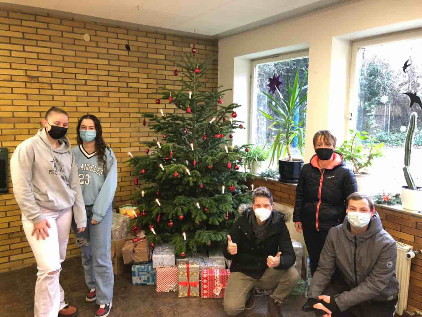 Fünf Personen mit Masken posieren lächelnd neben einem festlich geschmückten Weihnachtsbaum und zahlreichen Geschenken.