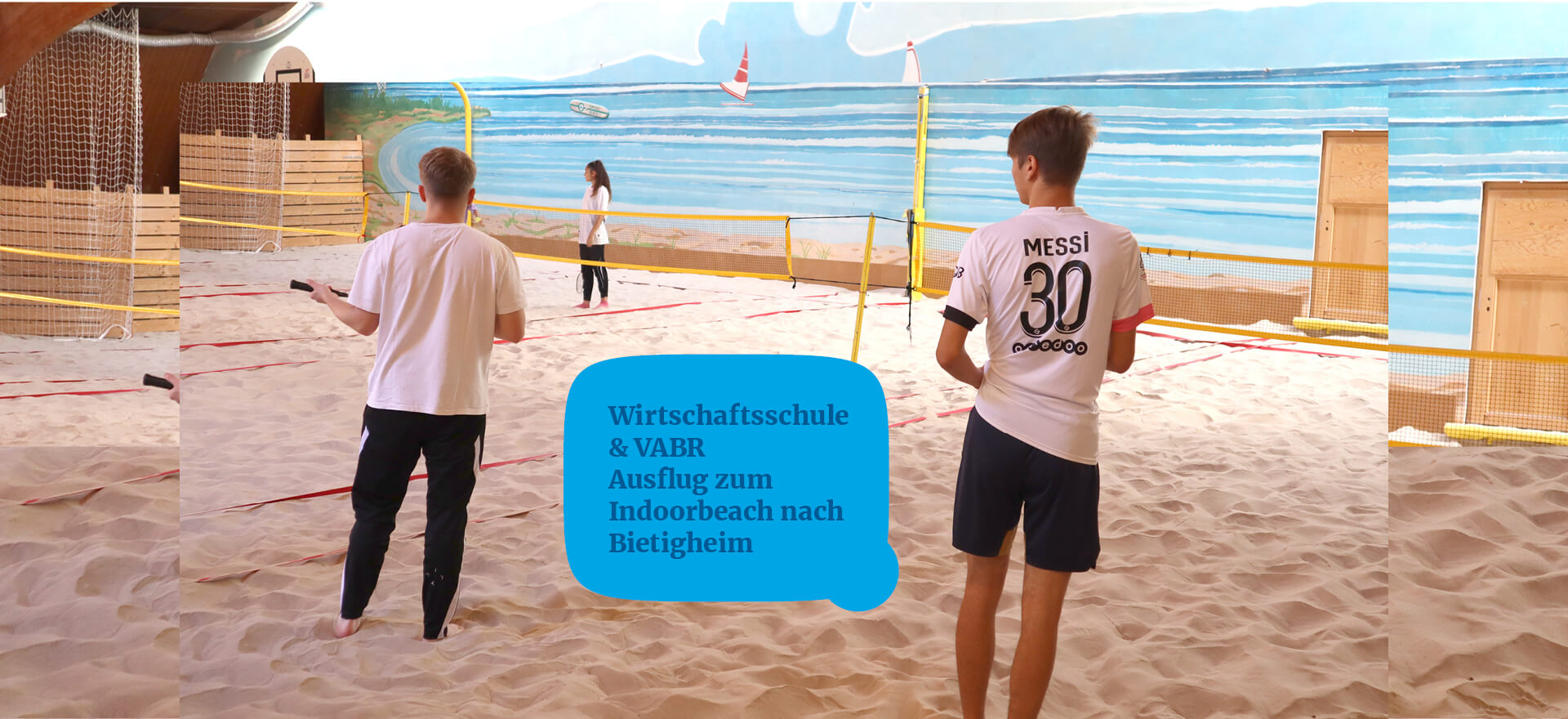 Zwei junge Männer spielen Beachvolleyball in einer Indoorhalle mit Sandboden und Strandmalerei an der Wand.