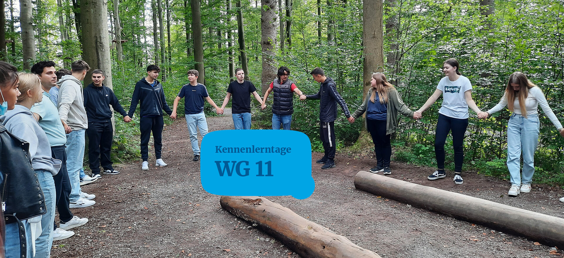 Eine Gruppe Jugendlicher steht im Wald im Kreis und hält sich bei einem Kennenlerntag an den Händen.