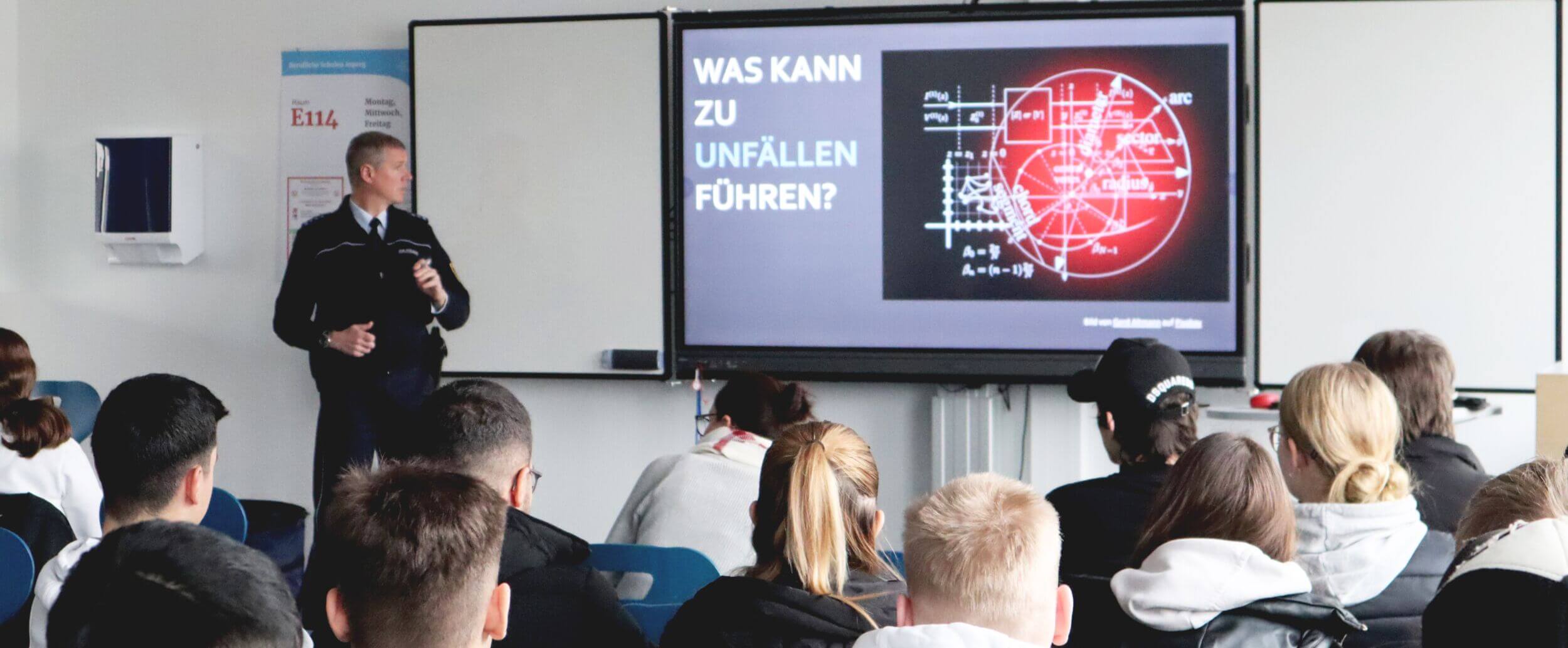 Ein Polizist hält vor einer Klasse einen Vortrag über das Thema - Was kann zu Unfällen führen? Die Präsentation ist auf dem Whiteboard zu sehen.