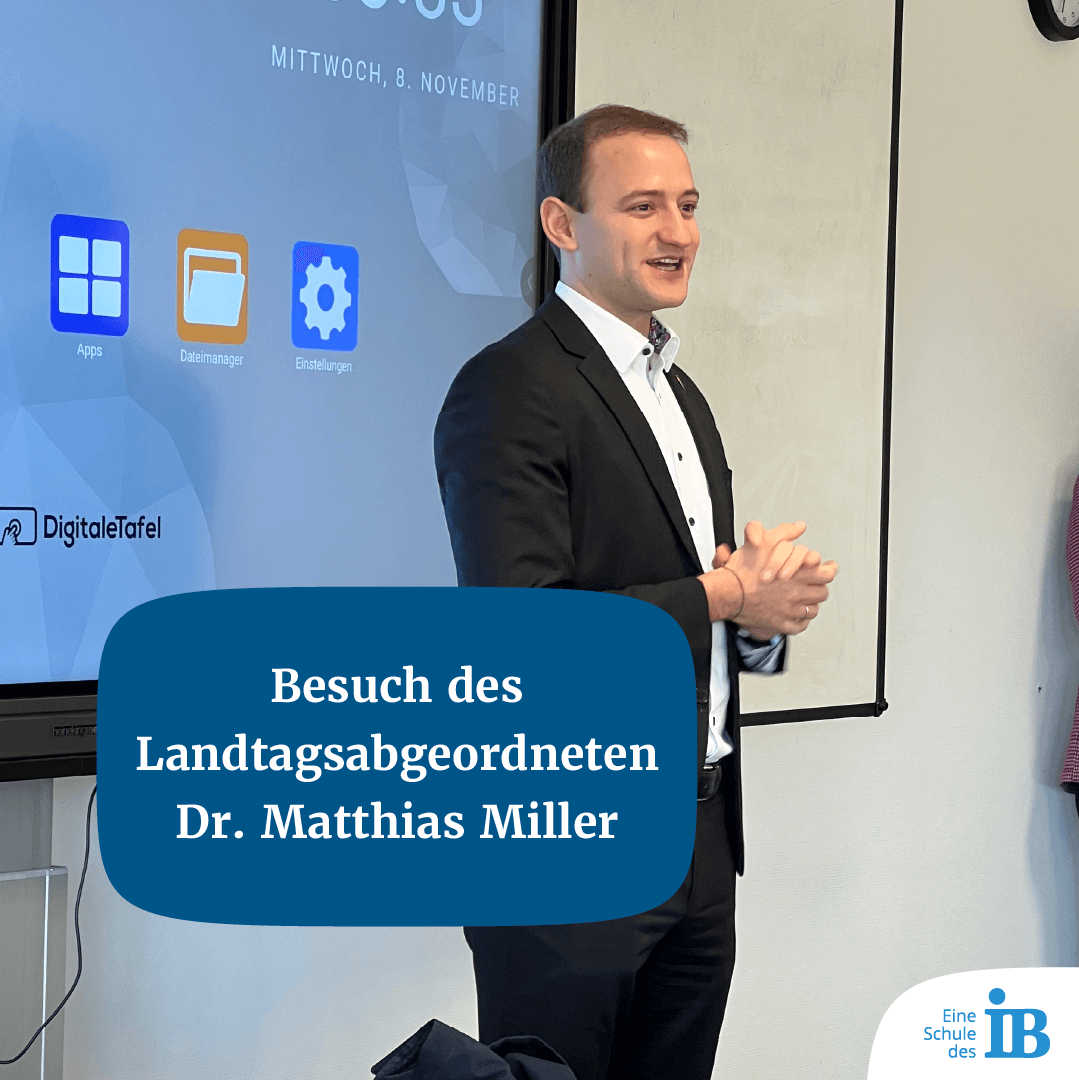 Ein Mann im Anzug hält einen Vortrag vor einer digitalen Tafel mit dem Thema "Besuch des Landtagsabgeordneten".