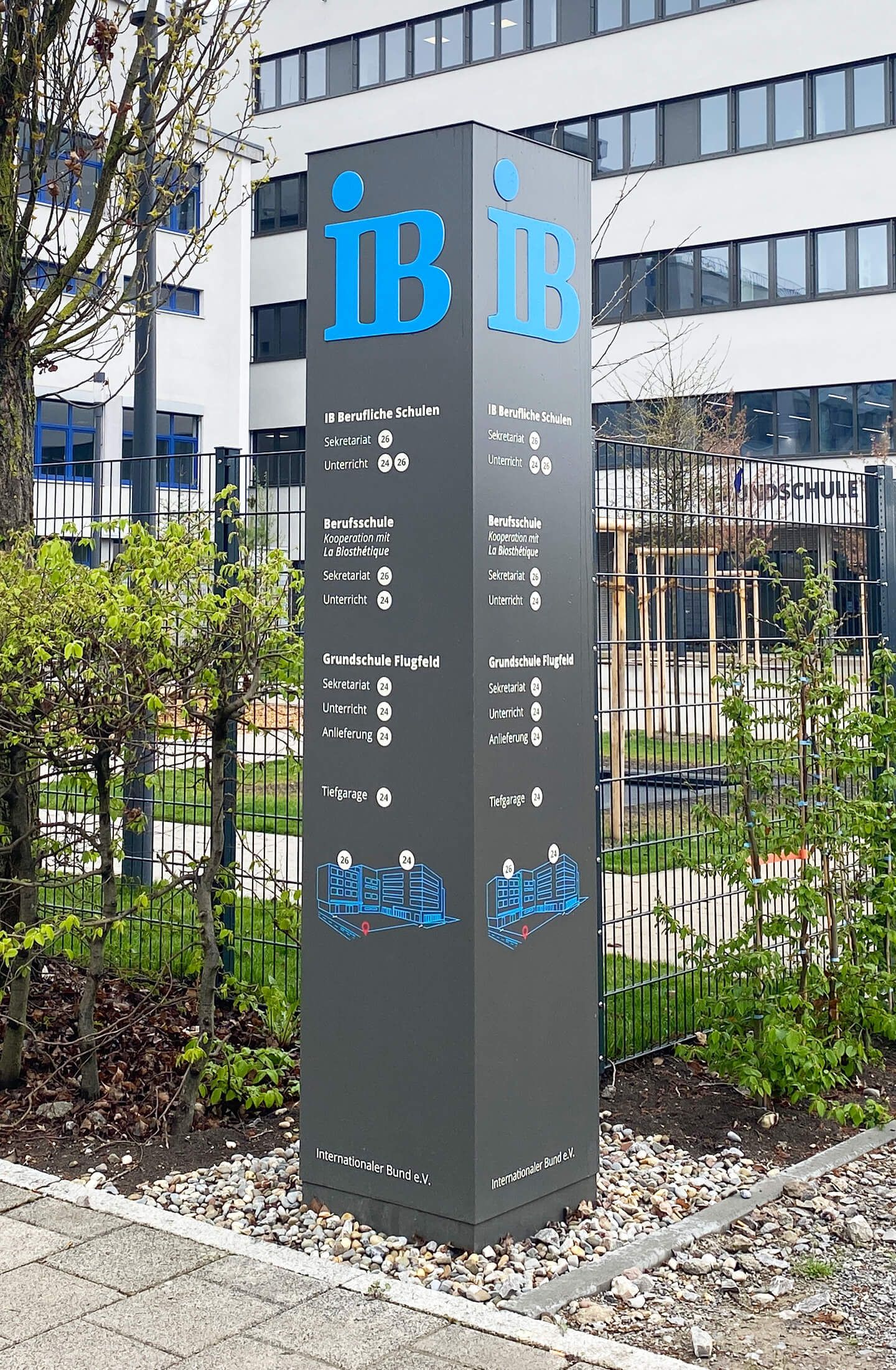 Ein Informationsstele des Internationalen Bundes zeigt die verschiedenen Bildungseinrichtungen im Außenbereich eines modernen Gebäudes.