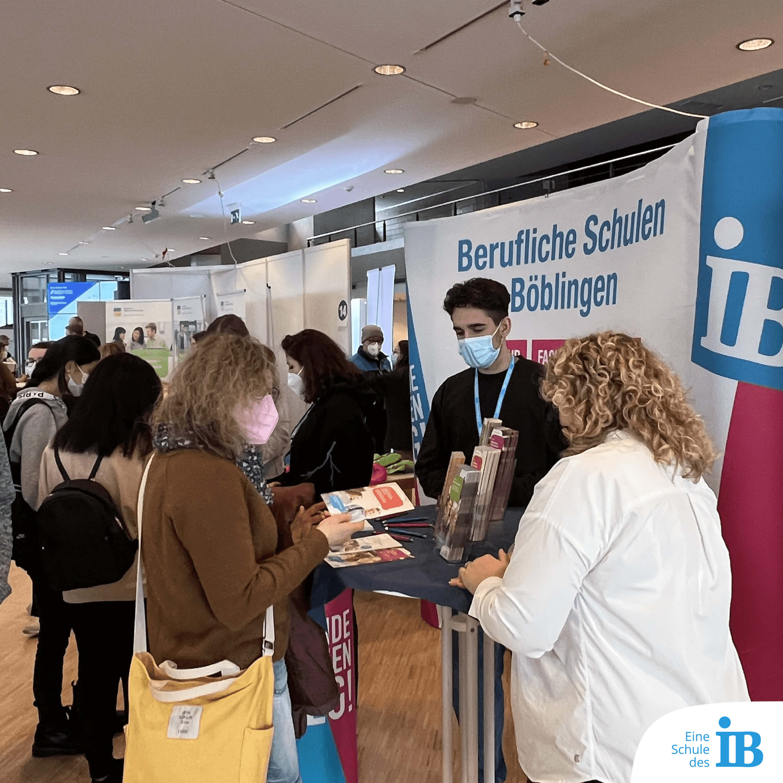 Messestand der IB Böblingen auf einer Berufsbildungsmesse - Eine Frau mit Maske informiert sich bei einer Lehrerin und einem Schüler.