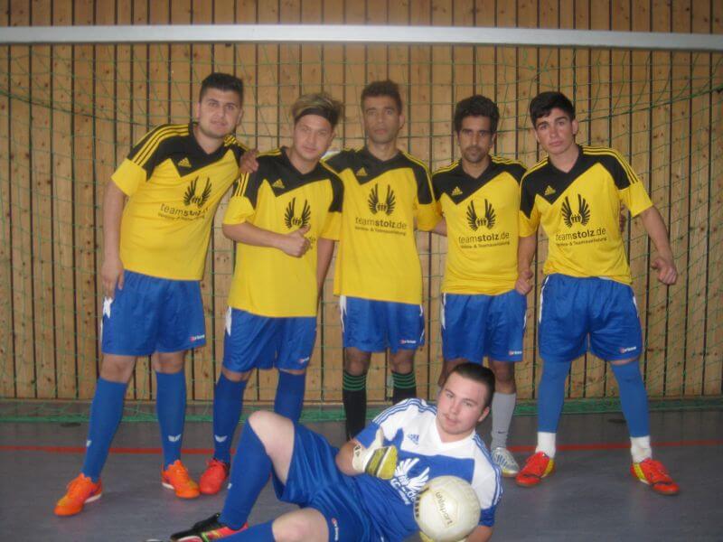 Eine Gruppe junger Männer in gelben Trikots posiert in einer Sporthalle mit einem liegenden Torwart und einem Fußball.
