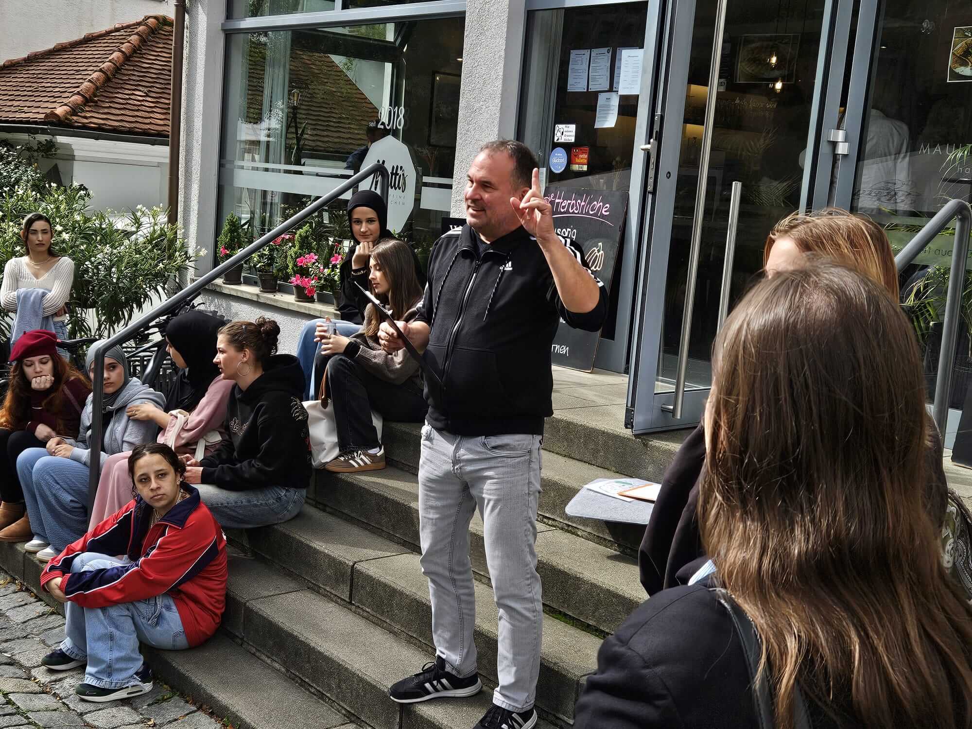 Eine Gruppe junger Menschen sitzt auf Treppenstufen vor einem Café, während ein Mann gestikulierend spricht.
