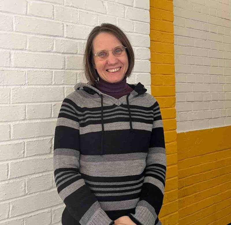 Eine lächelnde Frau in einem gestreiften Pullover steht vor einer weiß-gelben Ziegelwand.