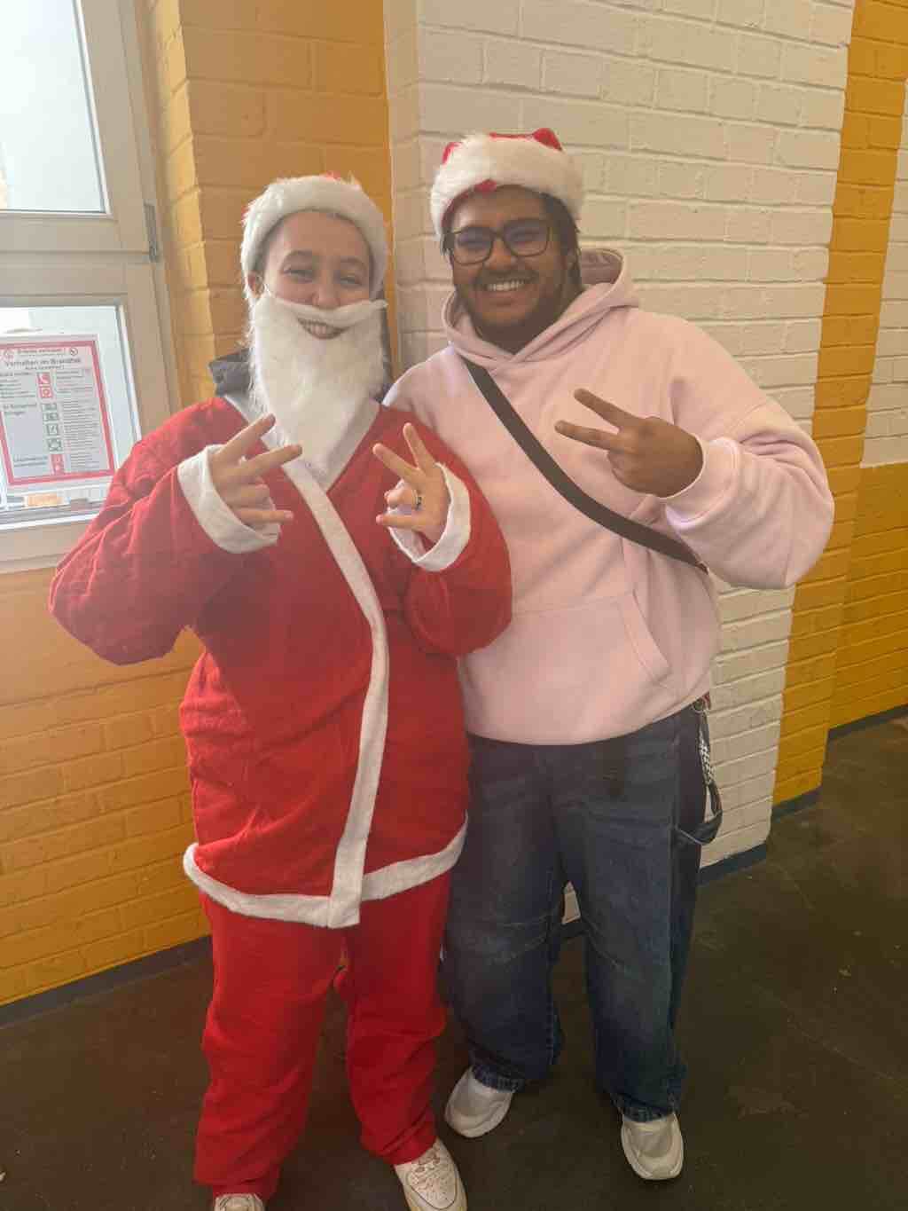 Zwei Personen in Weihnachtsmützen posieren fröhlich für ein Bild, wobei eine als Weihnachtsmann verkleidet ist.