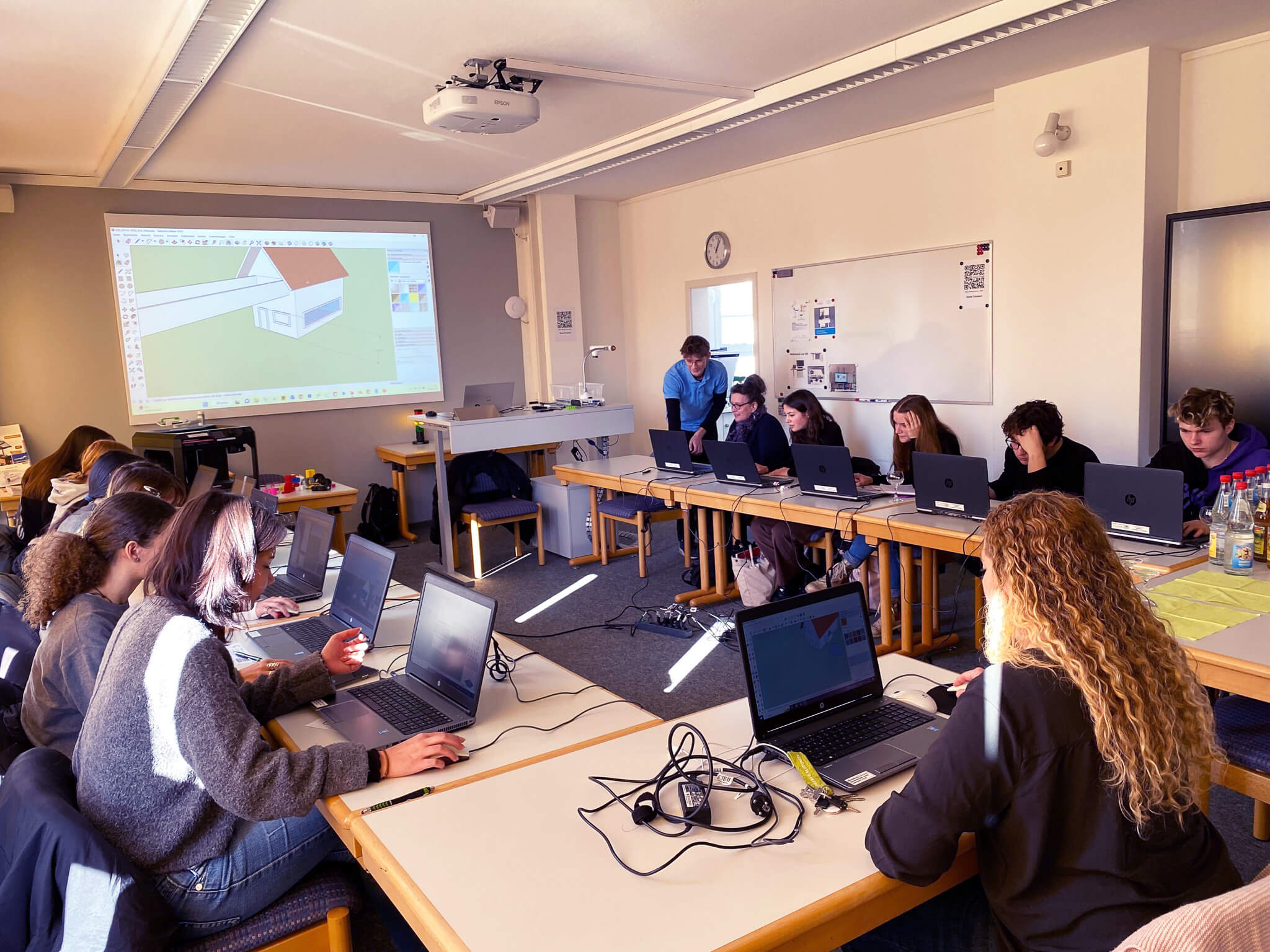 Eine Gruppe von Studierenden arbeitet an Laptops in einem Klassenraum, während ein Architekturmodell projiziert wird.