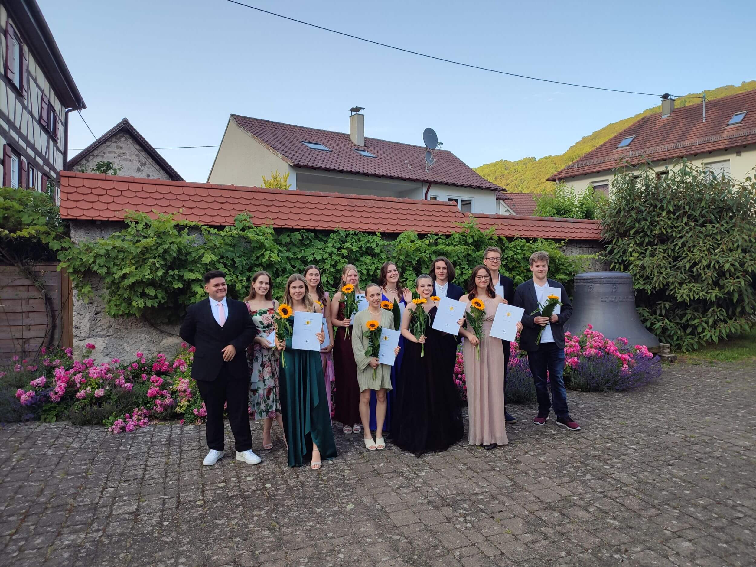 Eine Gruppe junger Menschen posiert draußen mit Urkunden und Sonnenblumen für ein Abschlussfoto.
