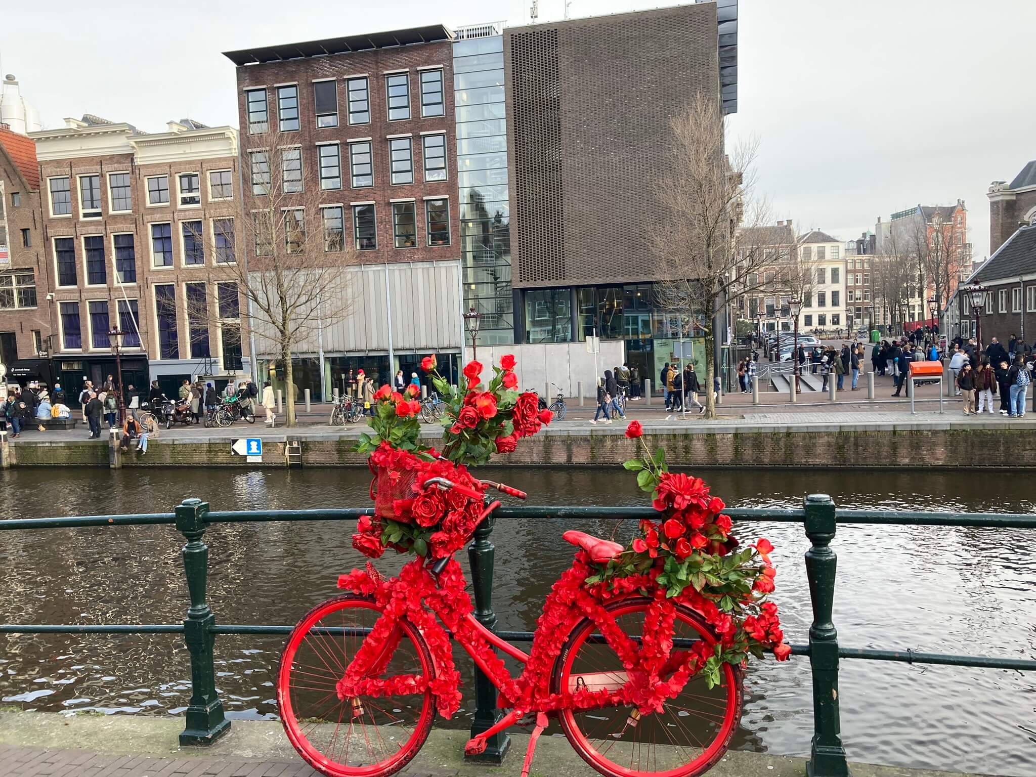 Ein mit roten Blumen geschmücktes Fahrrad steht an einem Kanal vor modernen und historischen Gebäuden.