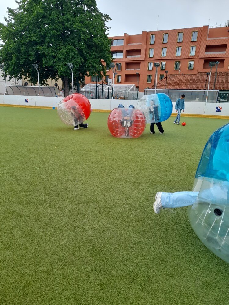 Kinder spielen in aufblasbaren Bällen auf einem Sportplatz, umgeben von Bäumen und Gebäuden im Hintergrund.