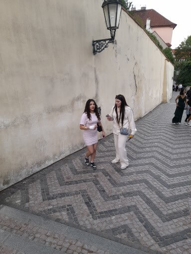 Zwei Frauen spazieren auf einem gepflasterten Weg entlang einer hellen Wand und unter einer Laterne.