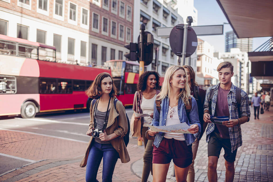 Eine Gruppe junger Menschen erkundet mit Karten eine belebte Stadt, während ein roter Doppeldeckerbus vorbeifährt.