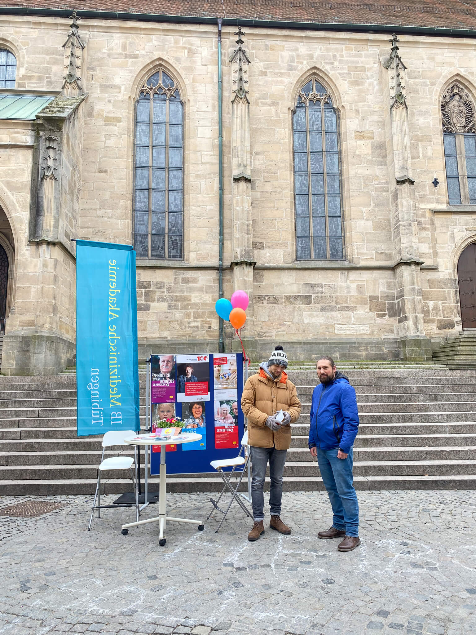 Zwei Männer stehen vor einem Infostand der Logopädieschule am Holzmarkt in Tübingen, mit bunten Luftballons dekoriert.