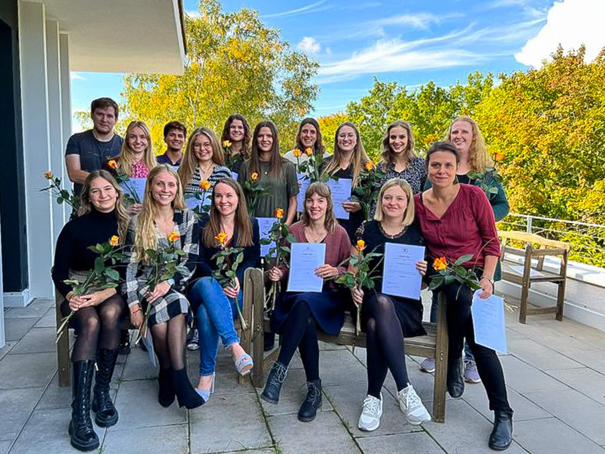 Eine Gruppe von Personen posiert mit Blumen und Zertifikaten für ein Abschlussfoto auf einer Terrasse.