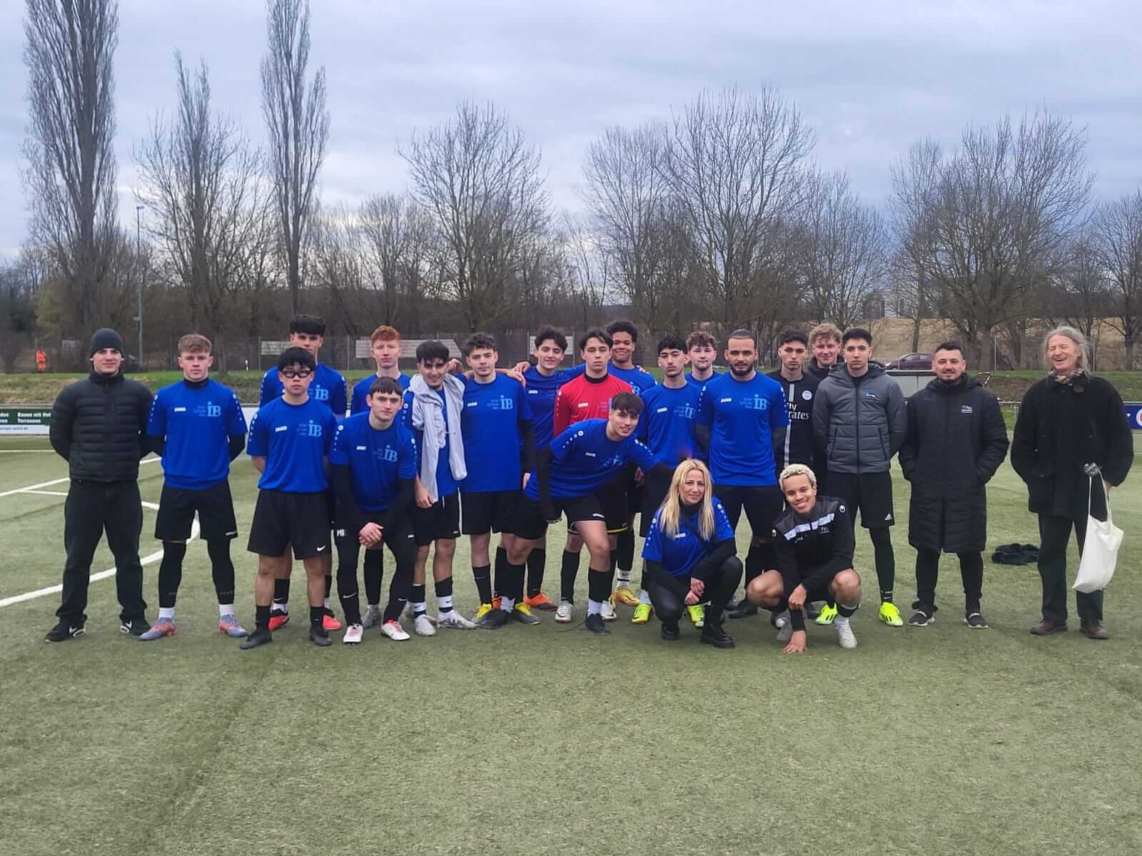 Eine Fußballmannschaft in blauen Trikots posiert gemeinsam auf einem grünen Spielfeld, umgeben von kahlen Bäumen.