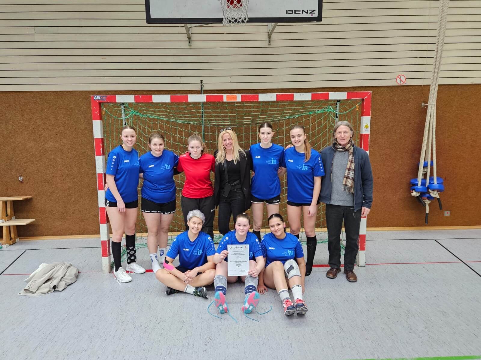 Eine Gruppe von Handballspielerinnen posiert in blauen Trikots vor einem Tor in einer Sporthalle mit einer Urkunde.