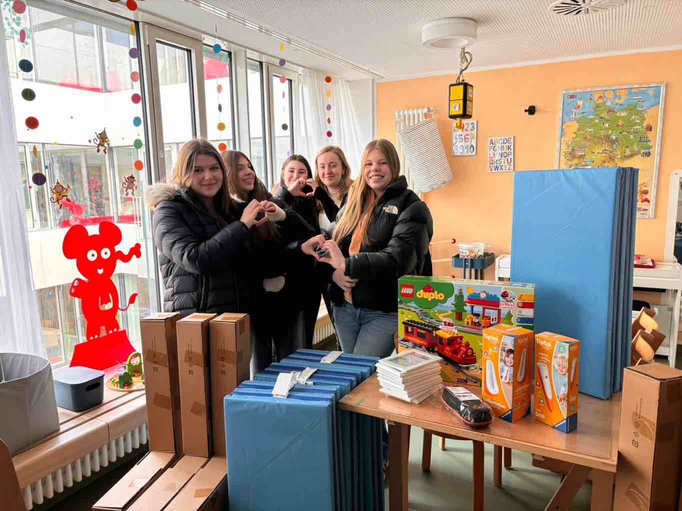 Fünf lächelnde Jugendliche stehen in einem farbenfrohen Raum neben Spielzeug und basteln ein Herz mit den Händen.