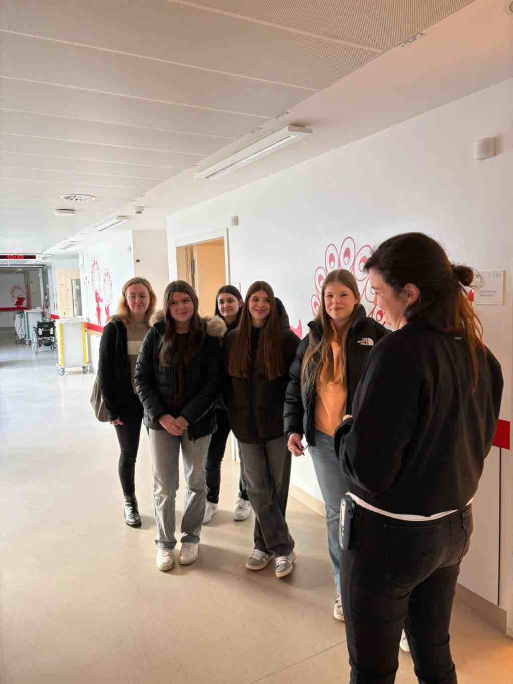 Eine Gruppe junger Frauen steht in einem hellen Flur und spricht mit einer Person in schwarzer Kleidung.