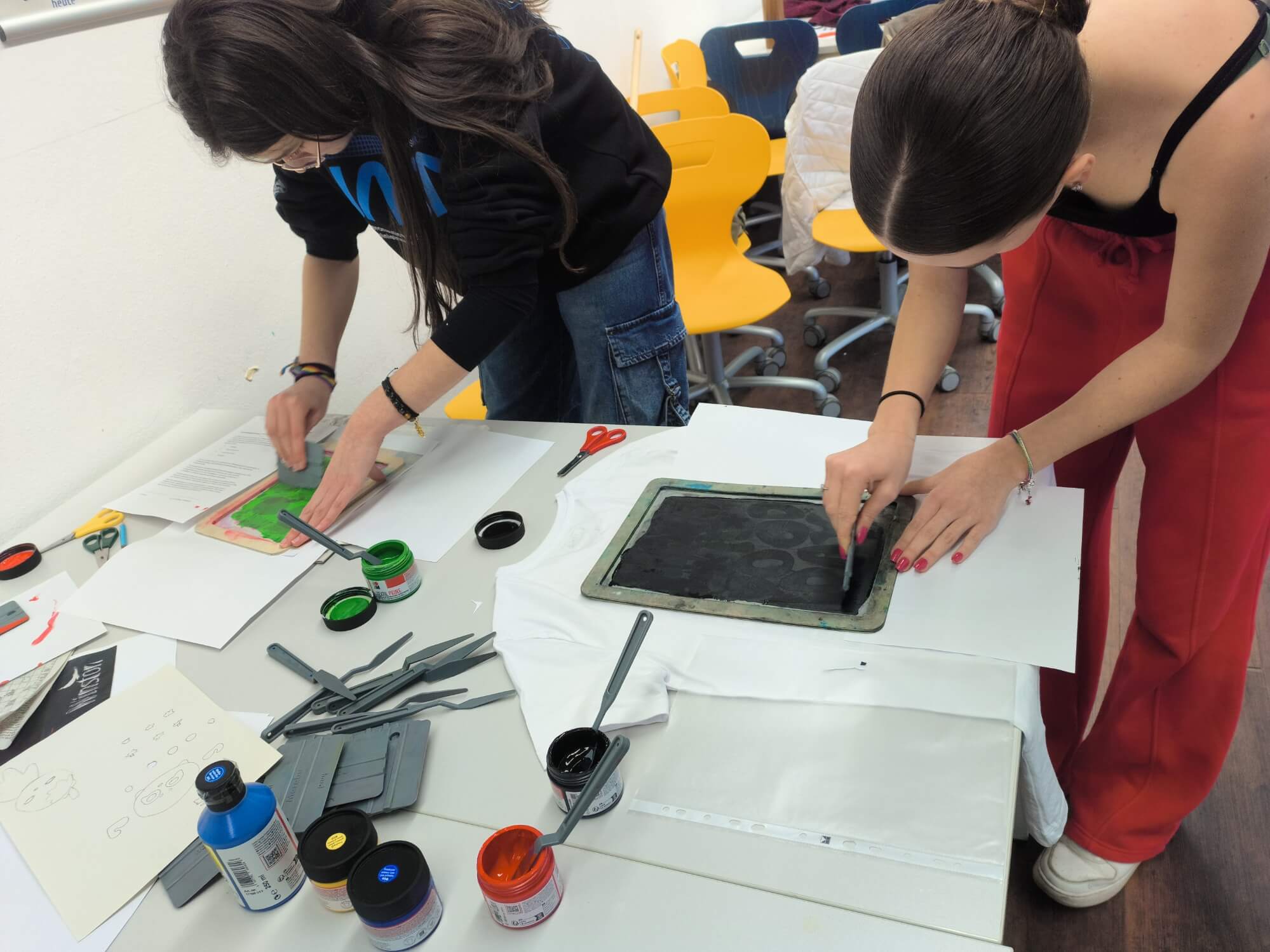 Zwei Schülerinnen arbeiten an einem Tisch und drucken mit Farben auf ein T-Shirt in einem kreativen Workshop.