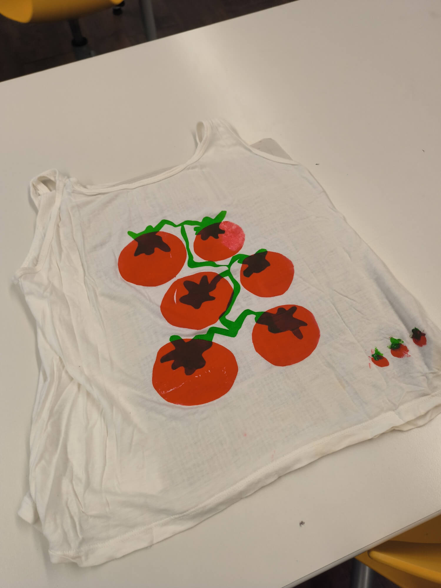 Ein weißes Oberteil mit bunten Tomatenmotiven liegt auf einem Tisch und zeigt kreative Handarbeitskunst.