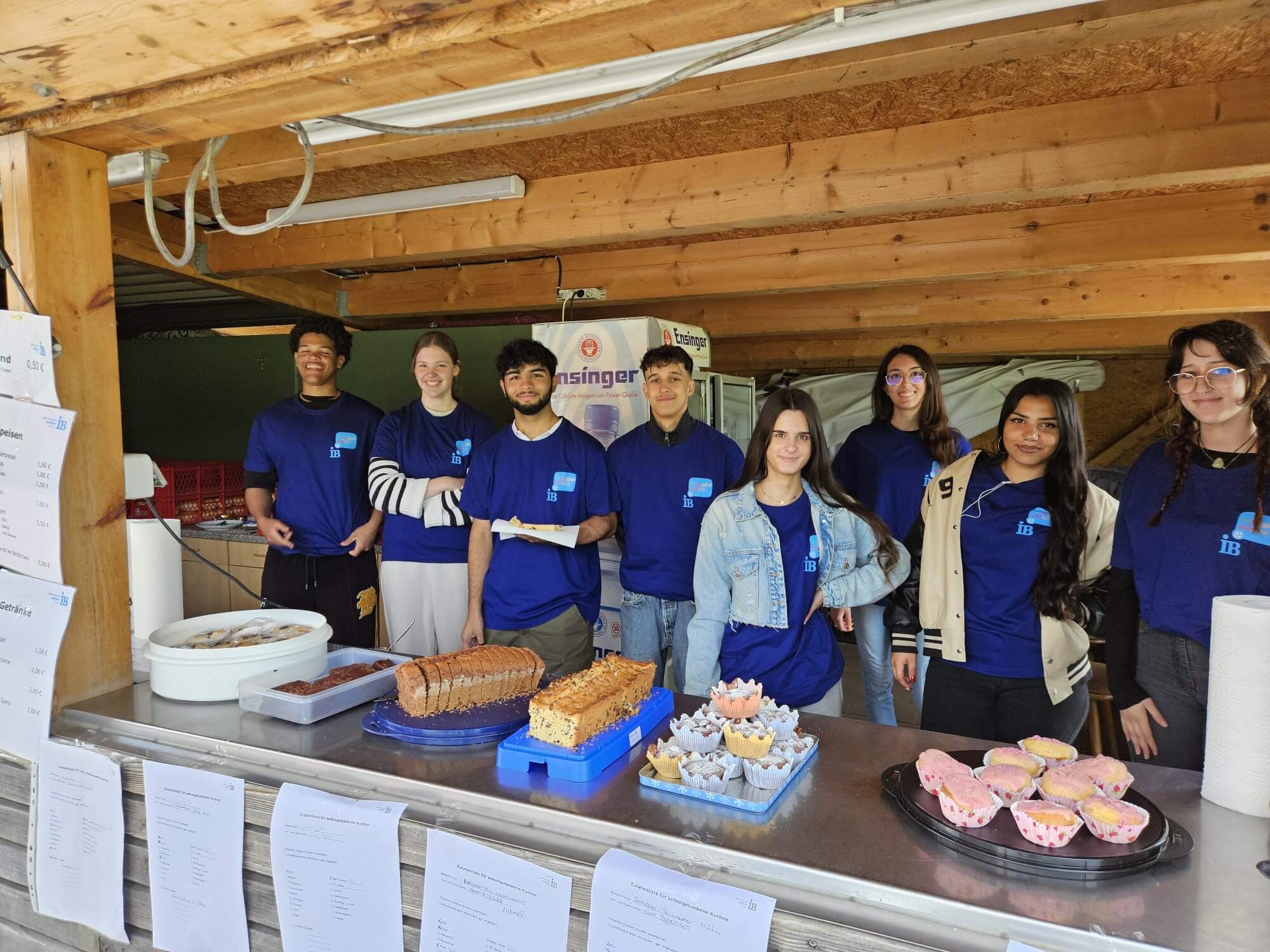 Eine Gruppe von acht jungen Menschen verkauft Kuchen und Muffins in einem hölzernen Verkaufsstand mit freundlichen Gesichtern.
