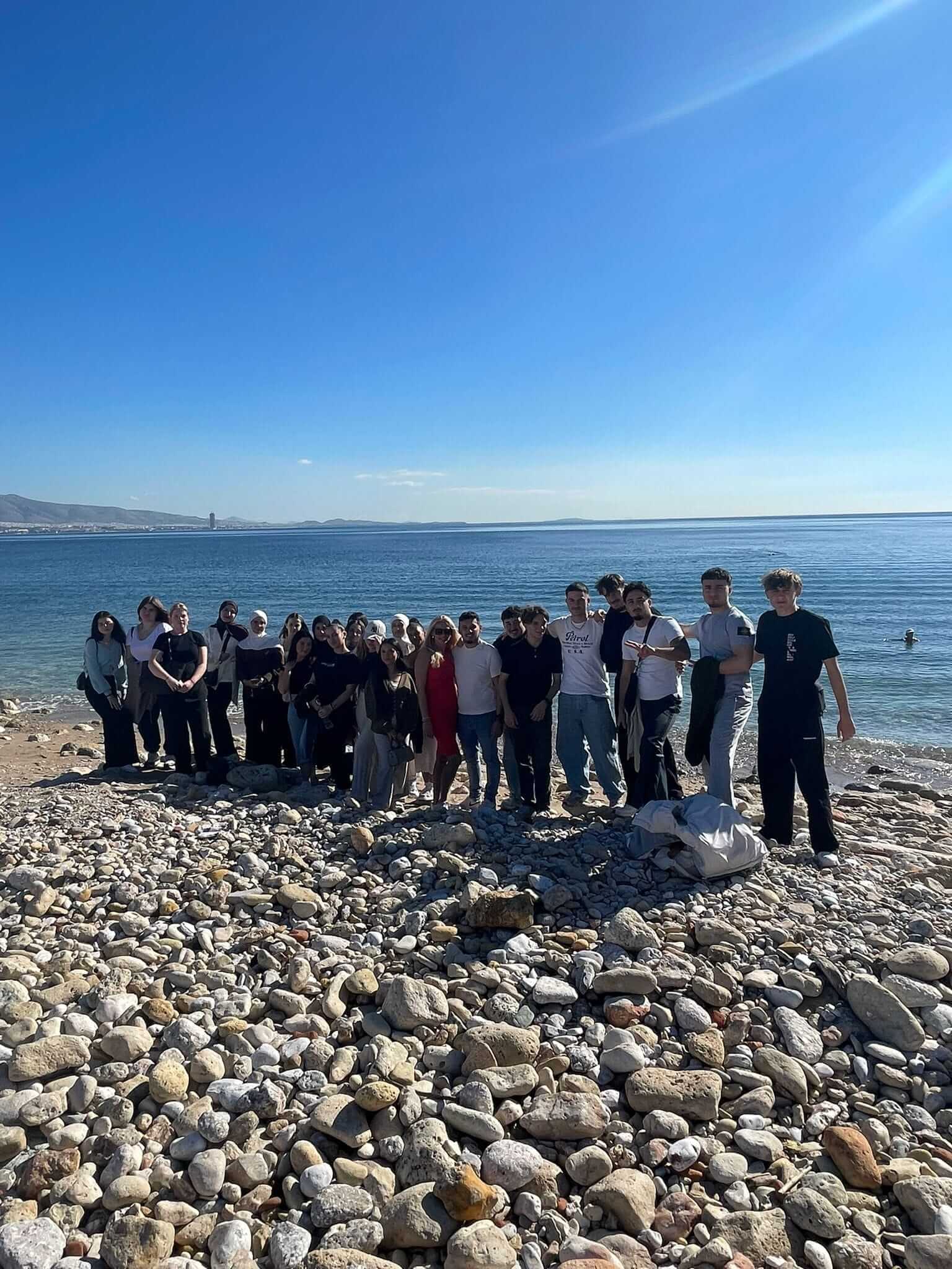 Eine Gruppe von Menschen versammelt sich an einem sonnigen Tag am steinigen Strand mit Blick auf das klare Meer.