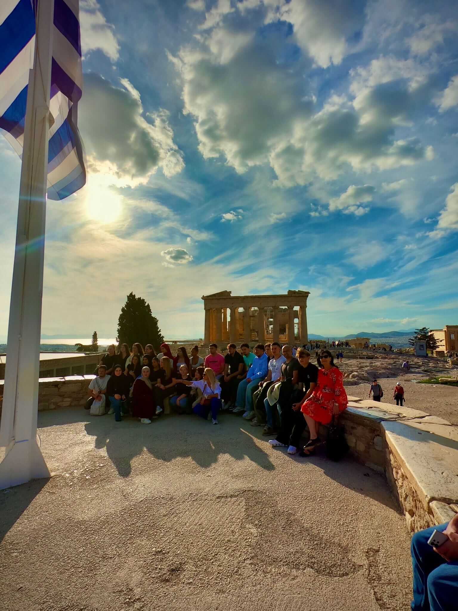 Eine Gruppe von Menschen sitzt vor dem Erechtheion in Athen, während die Sonne durch eine Wolkendecke scheint.