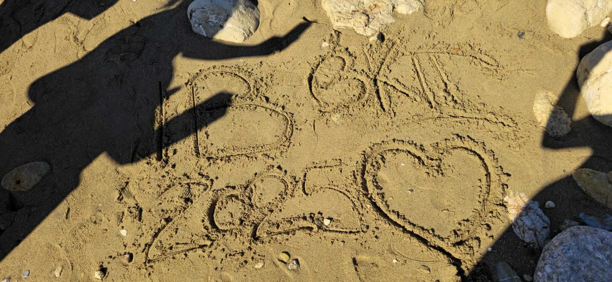 Im Sand sind die Buchstaben "B OKT 2025" und ein Herz mit Schatten von Menschen auf der Oberfläche gezeichnet.