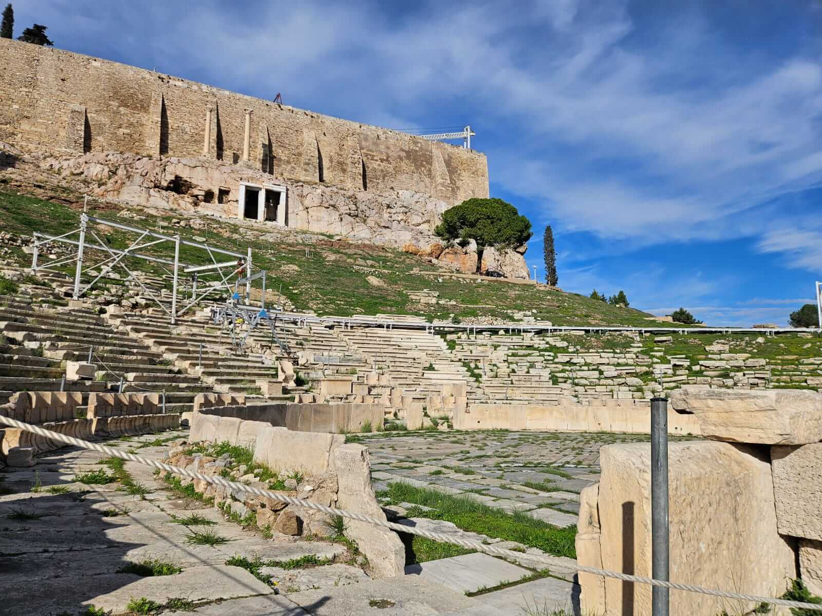 Das antike Theater mit steinernen Rängen liegt unter einem klaren blauen Himmel, umgeben von grüner Vegetation.