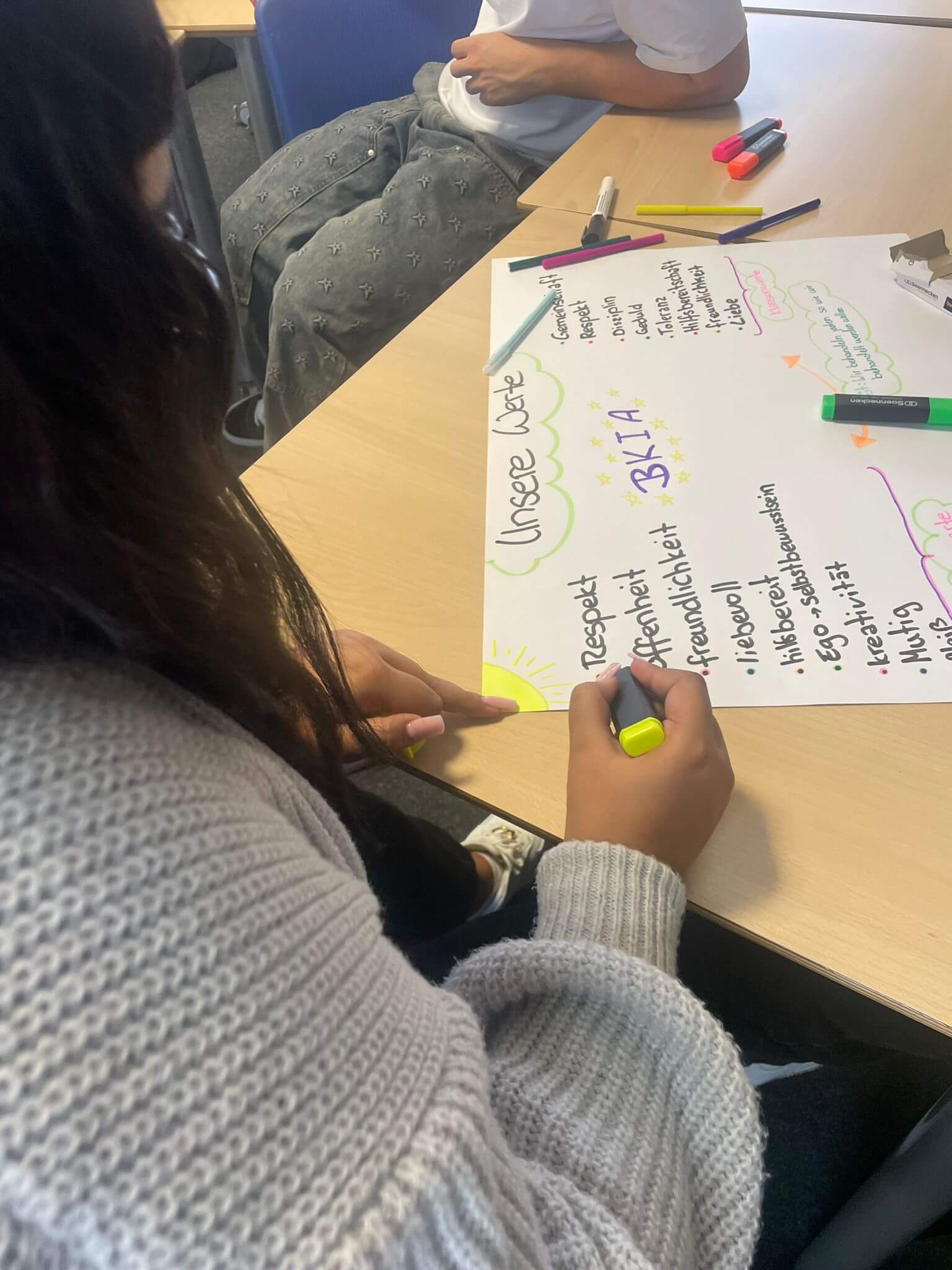 Eine Person schreibt mit einem gelben Stift auf ein Plakat mit dem Titel "Unsere Werte" in einem Klassenzimmer.