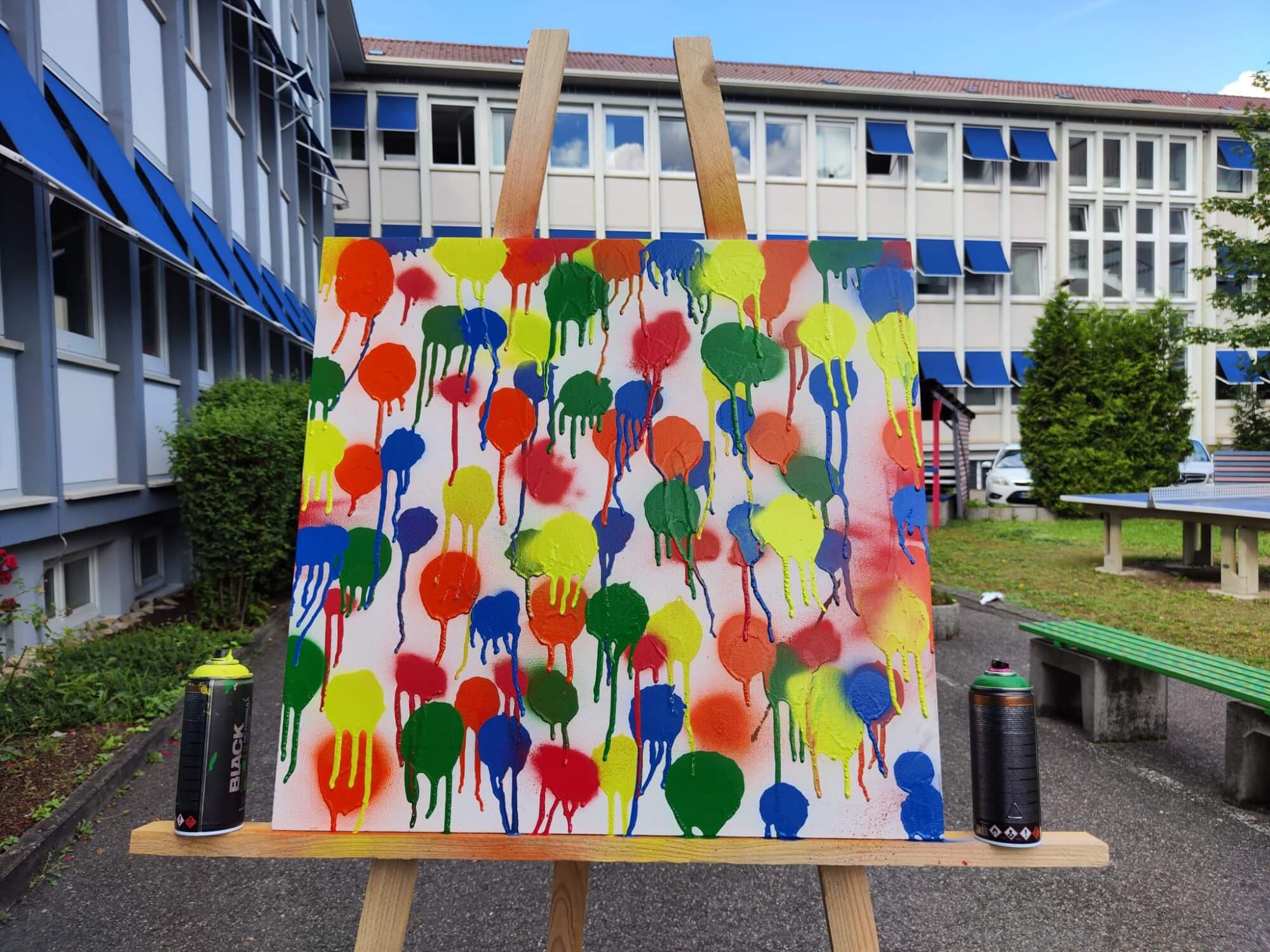 Ein buntes Graffiti-Kunstwerk mit Farbflecken steht auf einer Staffelei vor einem Schulgebäude im Freien.