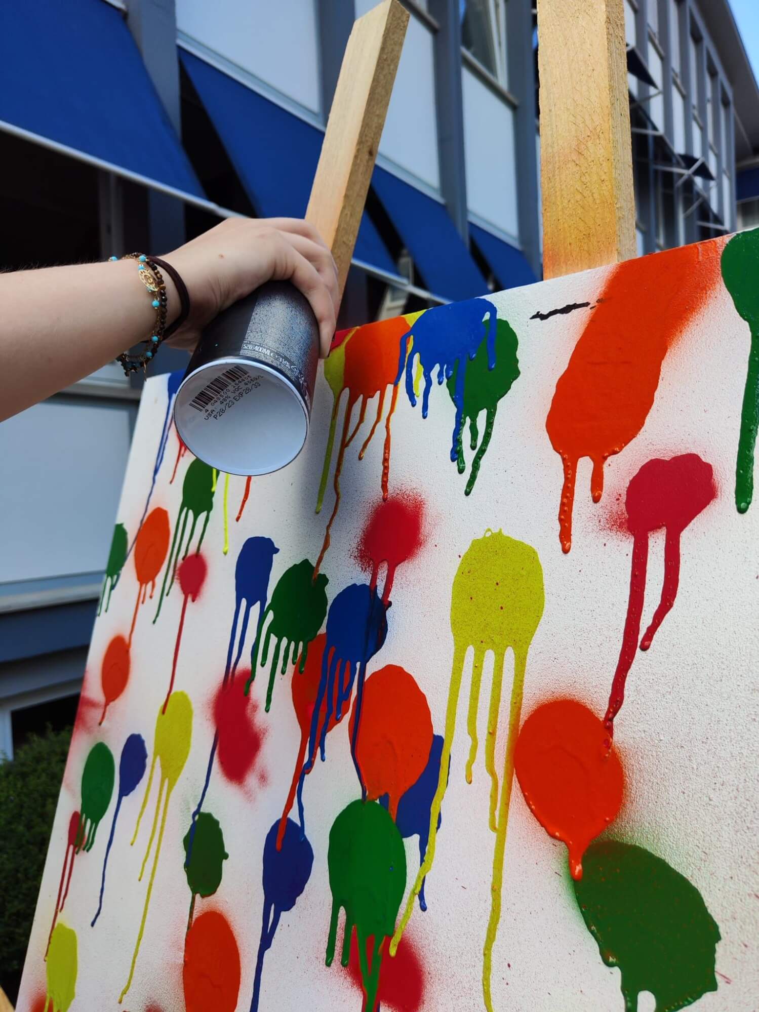Eine Hand sprüht bunte, tropfende Farbe auf eine Leinwand vor einem Gebäude mit blauen Markisen.