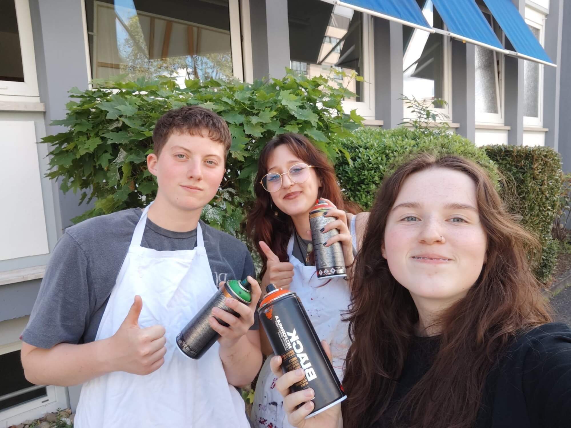 Drei junge Menschen posieren lächelnd mit Spraydosen vor einem Gebäude, bereit für ein kreatives Kunstprojekt im Freien.