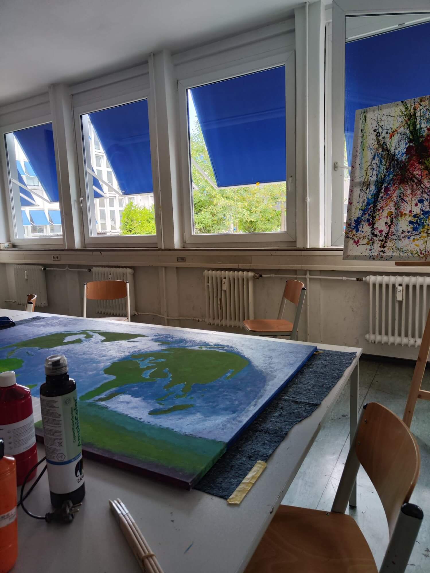 In einem Klassenzimmer mit blauen Jalousien liegt eine Weltkarte auf einem Tisch neben bunten Farbtöpfen und Pinseln.