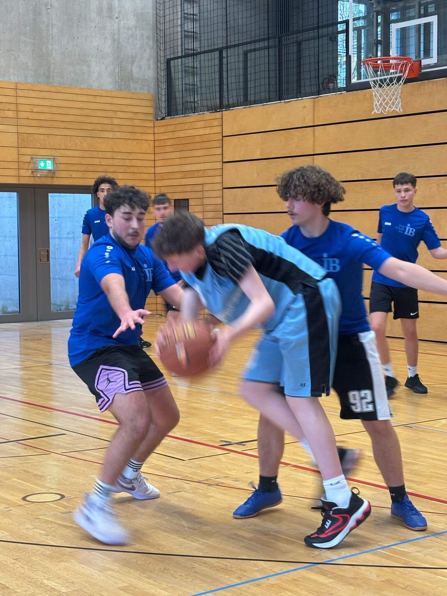 Zwei Basketballspieler in blauen Trikots versuchen, einem Gegenspieler in hellblauem Trikot den Ball abzunehmen.