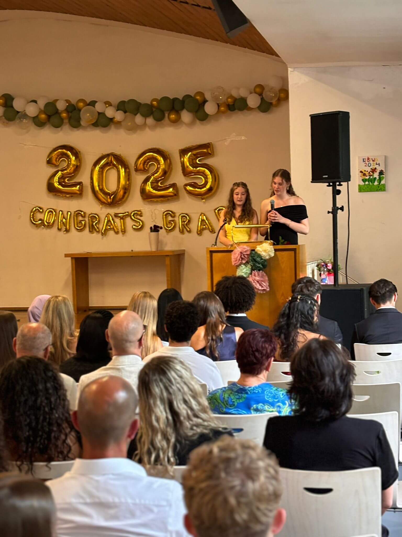 Zwei junge Frauen halten eine Rede auf einer Abschlussfeier im Jahr 2025, während das Publikum aufmerksam zuhört.