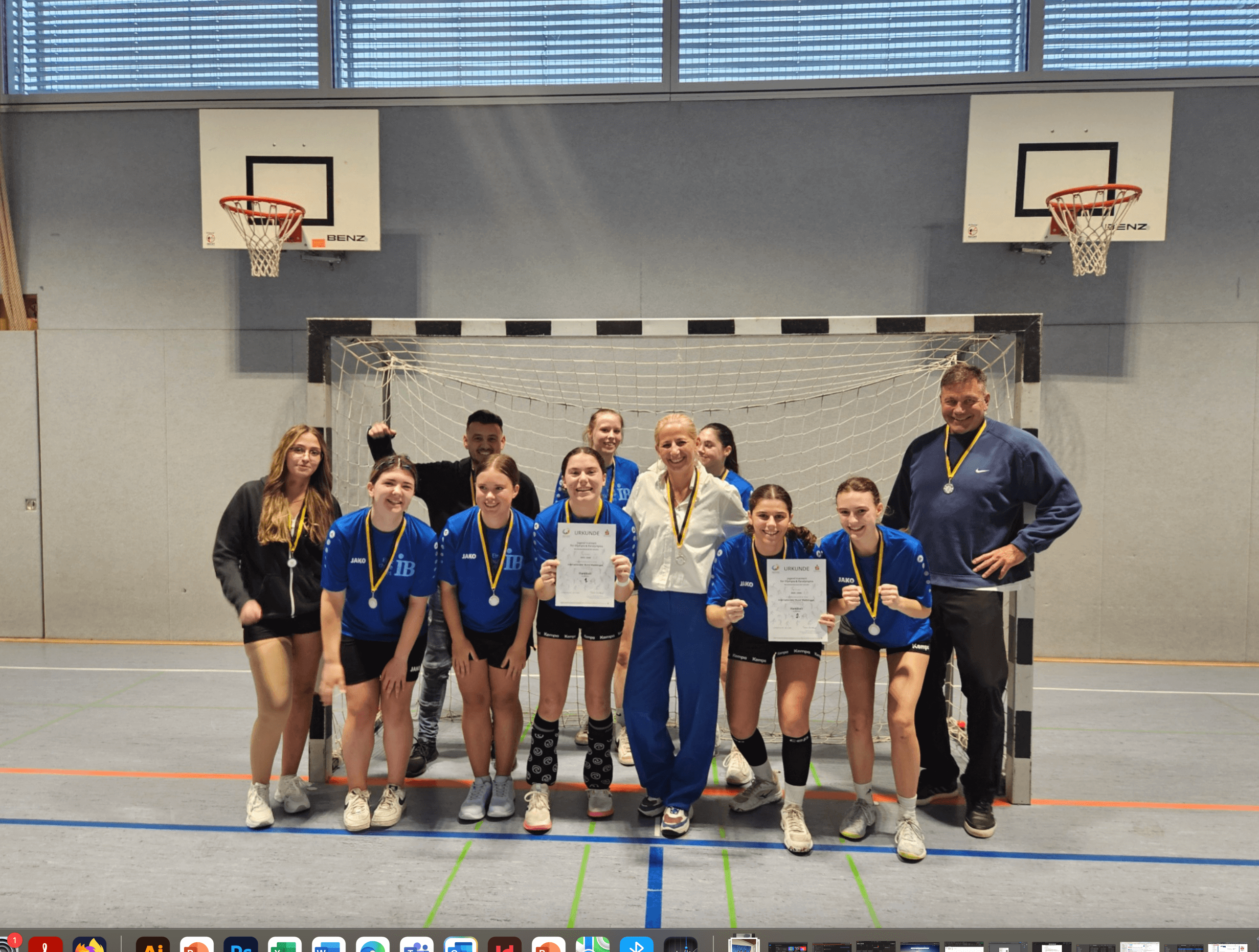 Eine Gruppe von jungen Athletinnen mit Medaillen posiert in einer Sporthalle vor einem Handballtor für ein Siegerfoto.