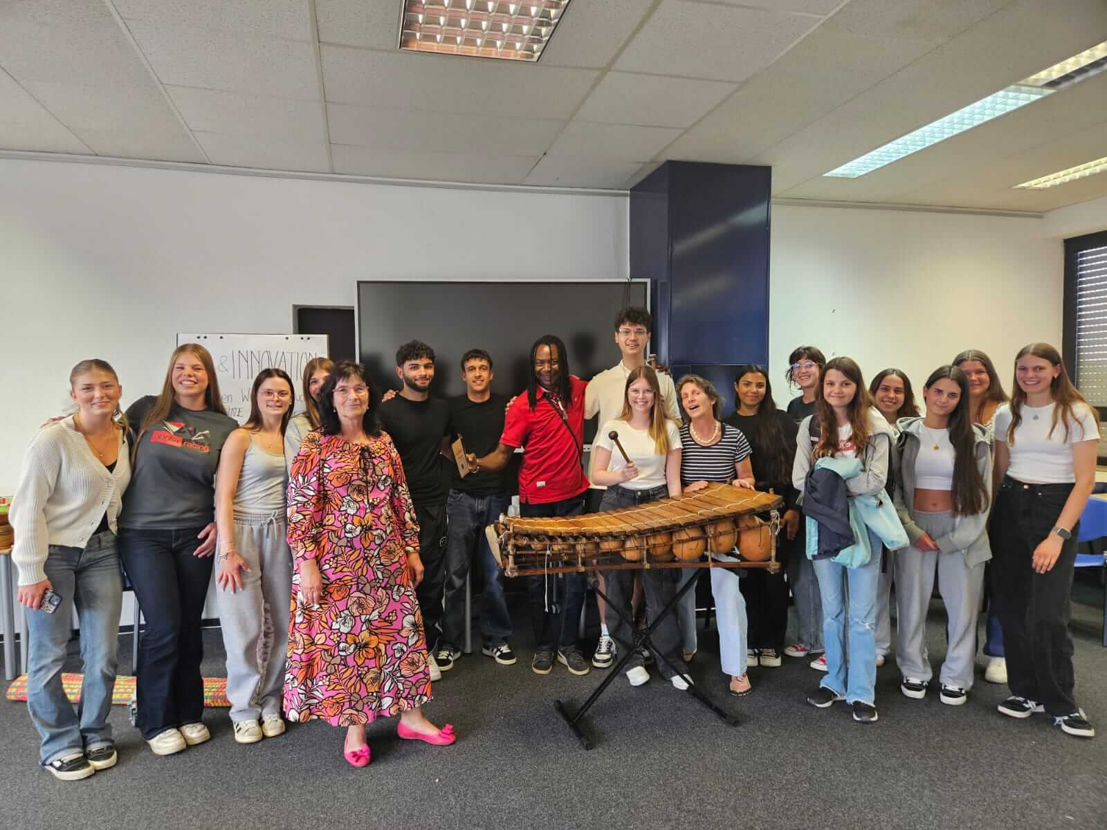 Eine Gruppe von Menschen posiert um ein traditionelles Xylophon in einem Raum, der für ein Treffen oder Workshop vorbereitet ist.