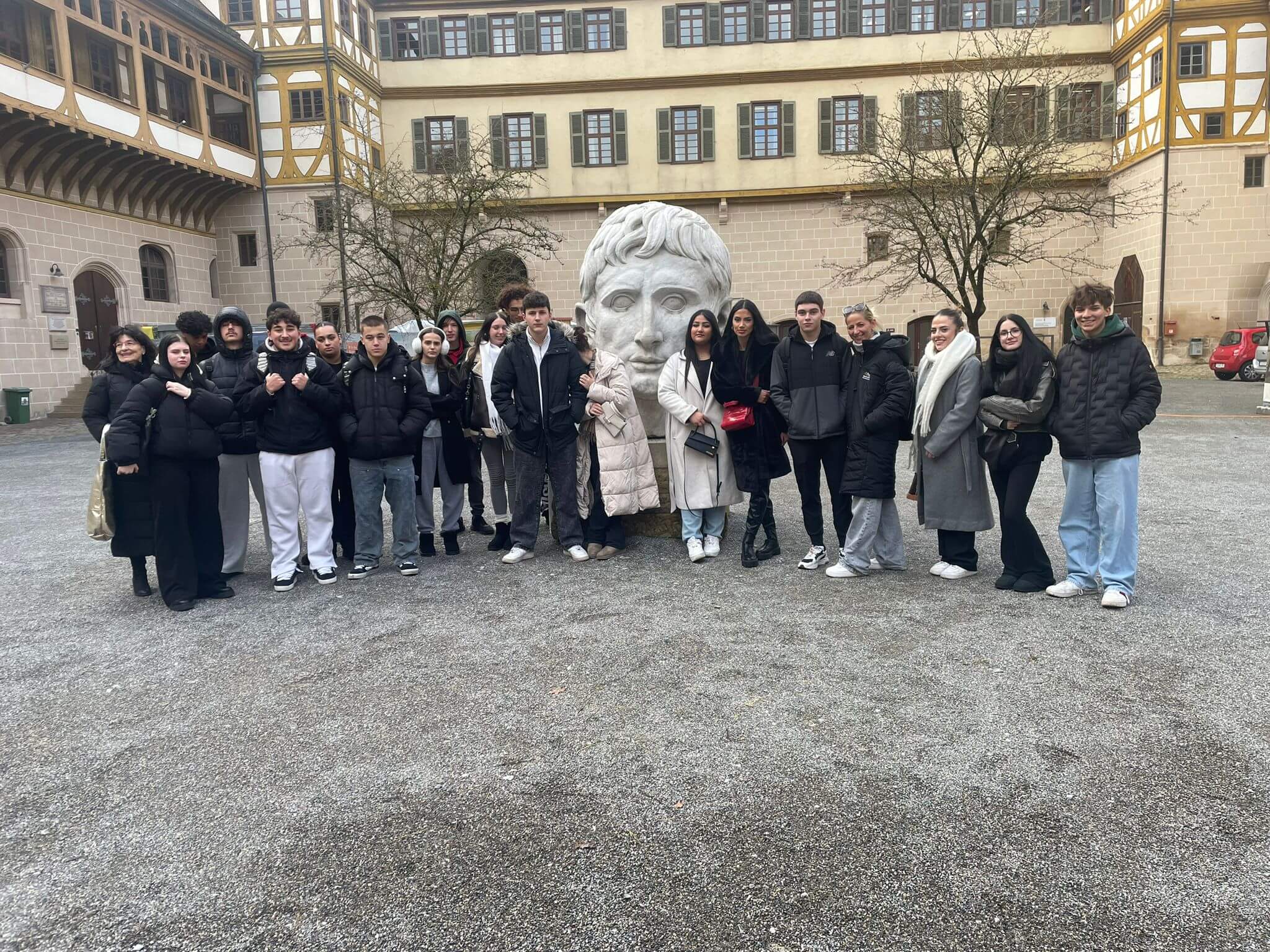 Eine Gruppe junger Menschen posiert vor einem großen Kopf-Skulptur auf einem historischen Innenhof.
