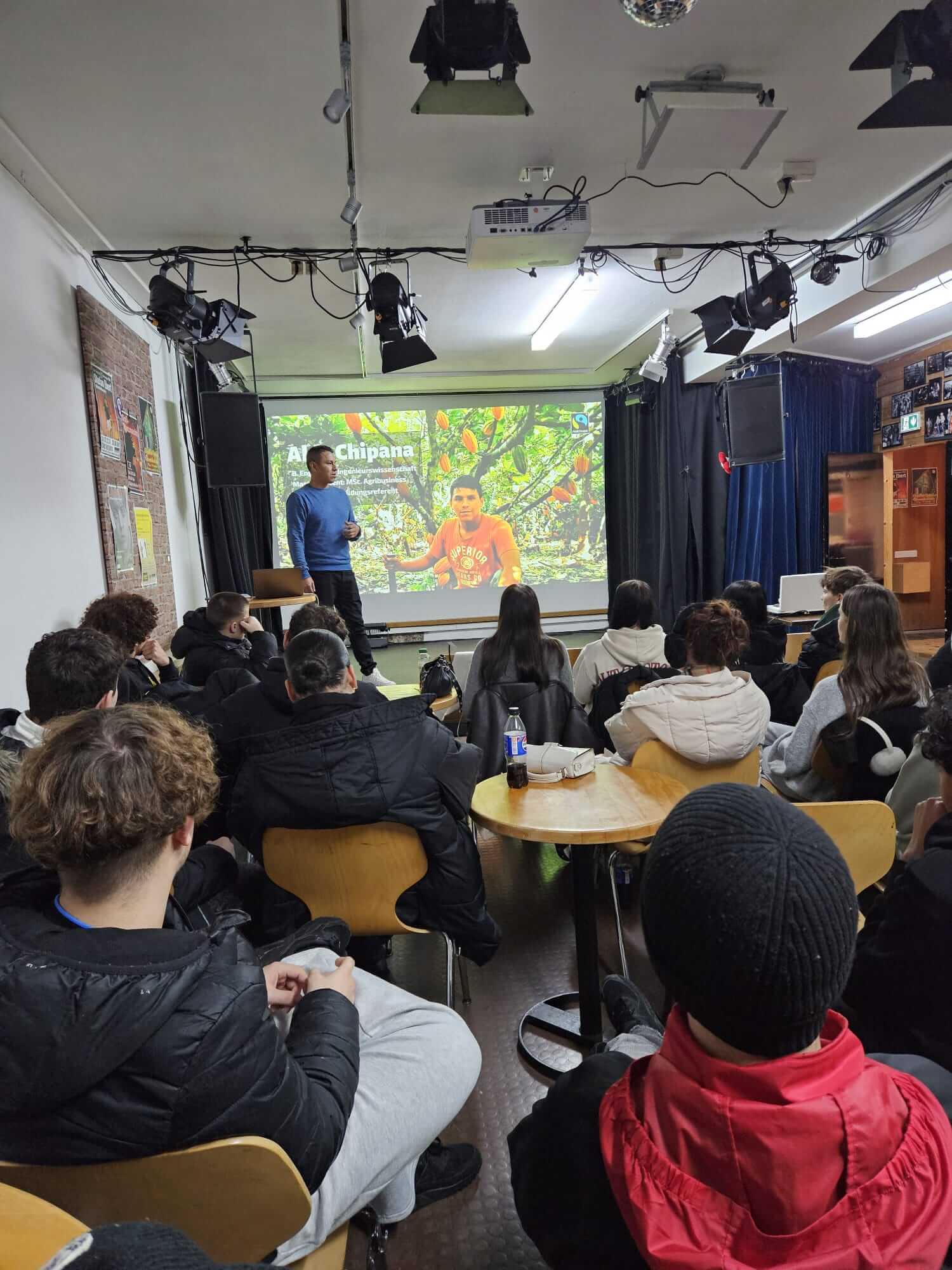 Ein Mann hält einen Vortrag vor einem Publikum in einem Raum, während ein Bild mit "Alan Chipana" auf der Leinwand projiziert wird.